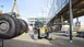 A yellow Liebherr telescopic loader is positioned on a construction site between a modern glass building and large tires being moved by a crane. In the background, construction cranes, scaffolding, and additional machinery are visible.