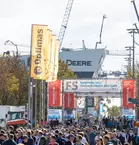 Crowd in the open-air site with machines, banners and cranes in the background. Large sign: 