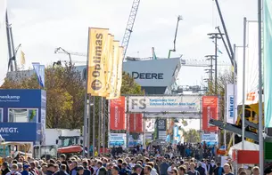 Crowd in the open-air site with machines, banners and cranes in the background. Large sign: 