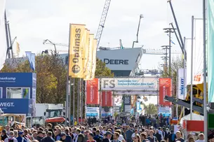 Crowd in the open-air site with machines, banners and cranes in the background. Large sign: 