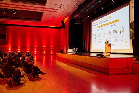 Ein Redner präsentiert auf einer Bühne mit großer Leinwand in einem rot beleuchteten Auditorium vor aufmerksam sitzendem Publikum.