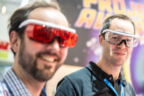 Two smiling people with colorful, modern-looking goggles in a technology exhibition.