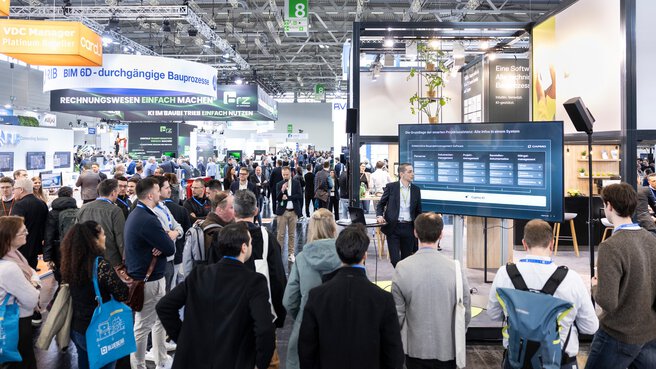 Trade show visitors are gathered around a screen, listening to a presentation at a booth.