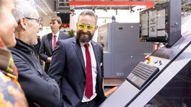 Mehrere Personen stehen an einer Maschine mit Display, Tastatur und rotem Notaus-Knopf. Ein Mann trägt eine neongelbe Brille.
