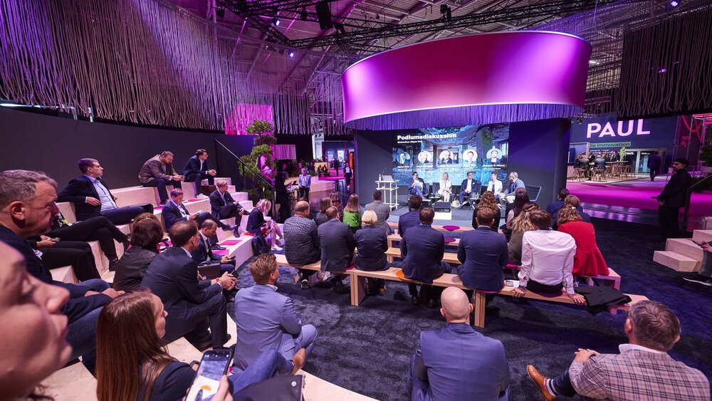 Interested visitors sit in a semicircle on wooden steps and benches and look towards a stage on which five people are holding a panel discussion.
