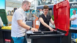 Two people examining a modern black waste container with a red lid and integrated sorting system at the Sulo trade fair stand.