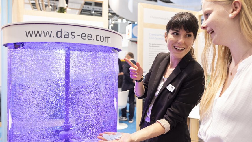 A woman in a black blazer demonstrates an exhibit with a purple glowing water tank full of air bubbles to a visitor at an exhibition stand.