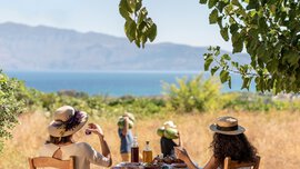 Two women are sitting at a table outside, enjoying food and drinks with a view of fields and the sea.