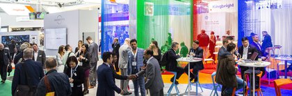 People bustle around in front of an exhibition stand with seating and colorful curtains.