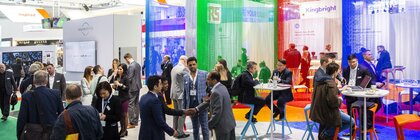 People bustle around in front of an exhibition stand with seating and colorful curtains.