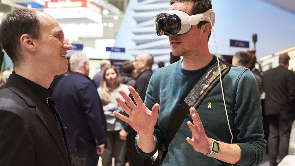 Two men are talking while one wears white VR glasses and gestures with his hands. Trade fair stands can be seen in the background.