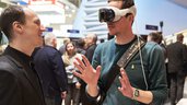 Two men are talking while one wears white VR glasses and gestures with his hands. Trade fair stands can be seen in the background.
