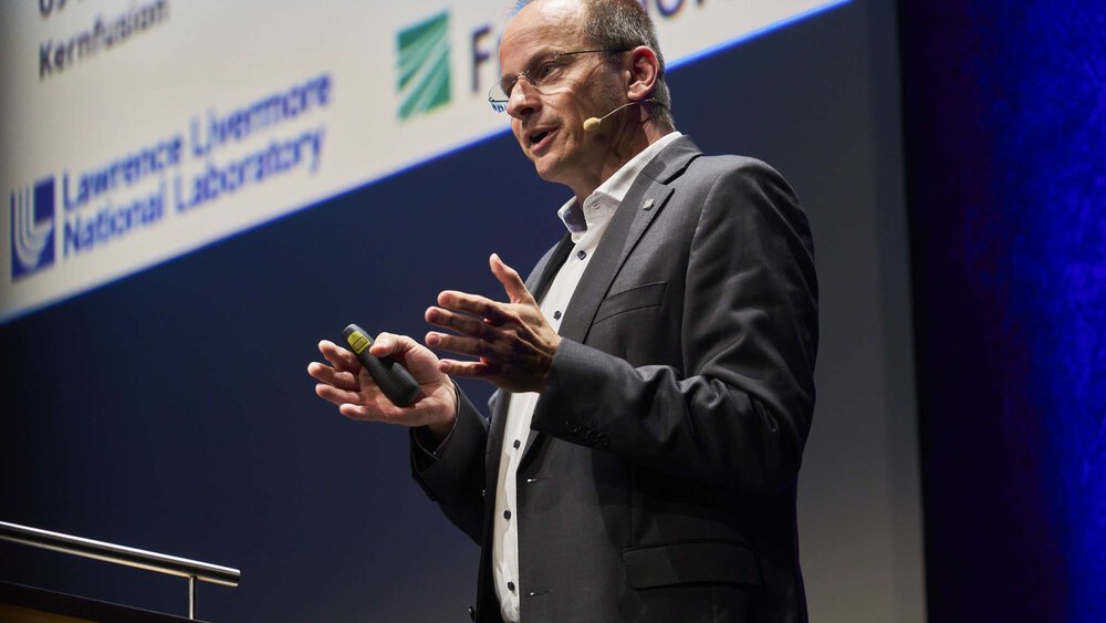 A man in a suit speaks on stage during a presentation and gestures with his hands.