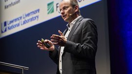A man in a suit speaks on stage during a presentation and gestures with his hands.