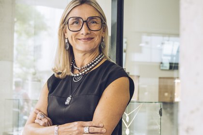 A smiling woman with shoulder-length hair and glasses wears several necklaces, earrings, bracelets and rings and has her arms crossed.