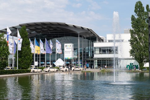 Exterior view of the Munich trade fair grounds showing the main entrance to IFAT, with a reflecting pool and fountain in the foreground, flagpoles bearing IFAT and partner flags, and several vehicles and visitors in front of the building.