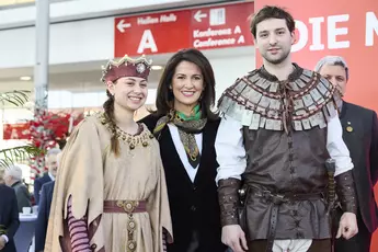 Three people in front of a stage at a trade fair opening ceremony. The people on the right and left are wearing traditional clothing from the Bavarian Forest, the trade fair's partner country.