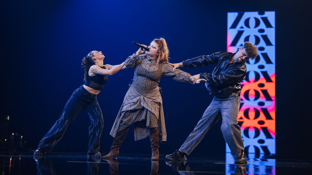 Three people on a stage, the woman in the centre sings into a microphone while a man and a woman pull her from both sides.