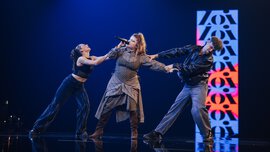 Three people on a stage, the woman in the centre sings into a microphone while a man and a woman pull her from both sides.