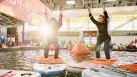 Zwei Personen in Neoprenanzügen üben Yoga-Posen auf Paddelbrettern in einem Hallenbad mit hellem Sonnenlicht im Rücken.