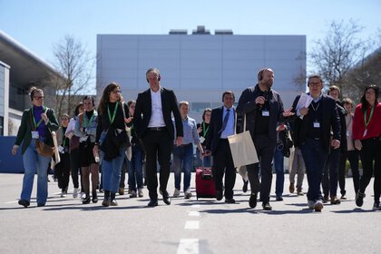 Eine Gruppe Messebesuchern mit Kopfhörern folgt bei einer Führung einem Mann mit Mikrofon, sie gehen auf einer breiten Straße unter blauem Himmel.