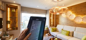 Eine Hand, die ein Tablet hält, steuert ein Smart-Home-System in einem modernen Wohnzimmer mit neutralen Tönen und stilvoller Beleuchtung.