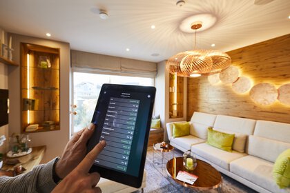 Eine Hand, die ein Tablet hält, steuert ein Smart-Home-System in einem modernen Wohnzimmer mit neutralen Tönen und stilvoller Beleuchtung.
