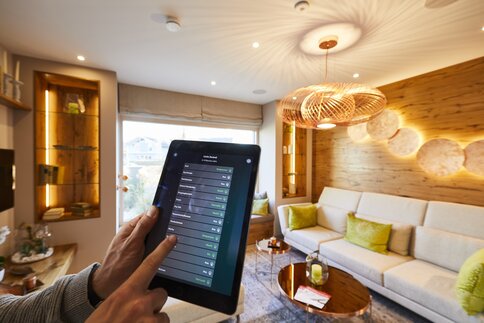 Eine Hand, die ein Tablet hält, steuert ein Smart-Home-System in einem modernen Wohnzimmer mit neutralen Tönen und stilvoller Beleuchtung.