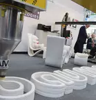 A 3D printer creates the bauma logo from concrete while two people are discussing in the background.