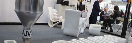 A 3D printer creates the bauma logo from concrete while two people are discussing in the background.