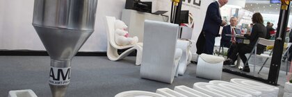 A 3D printer creates the bauma logo from concrete while two people are discussing in the background.