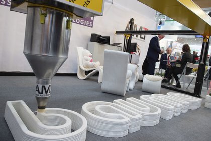 A 3D printer creates the bauma logo from concrete while two people are discussing in the background.