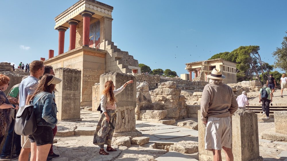 A tour guide explains the ruins of the Palace of Knossos to a group of tourists under a bright blue sky.