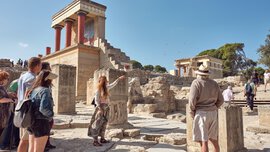 A tour guide explains the ruins of the Palace of Knossos to a group of tourists under a bright blue sky.