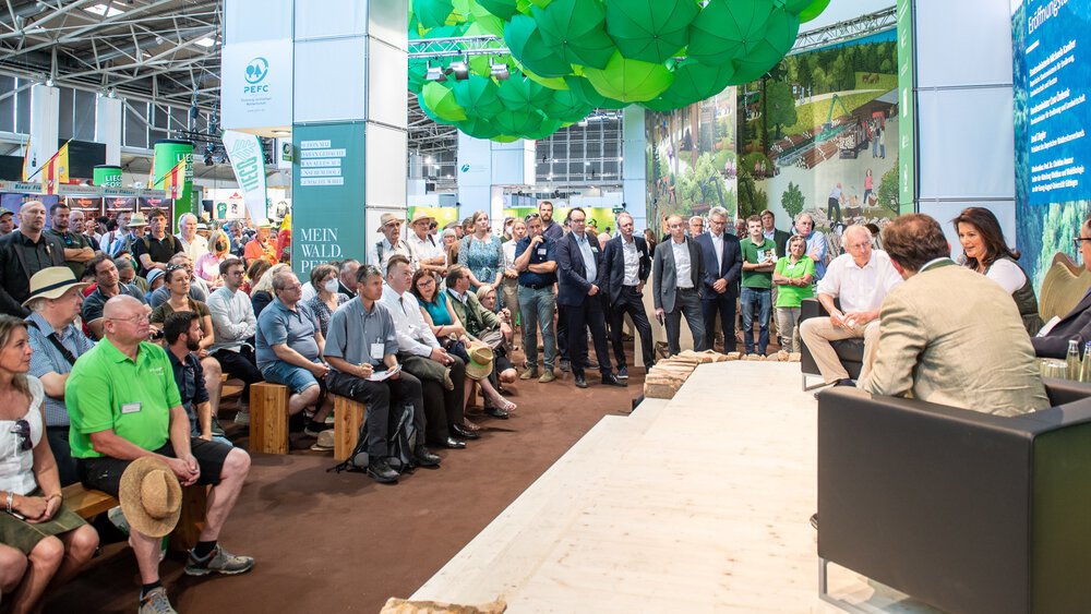 Großes Publikum verfolgt eine Podiumsdiskussion an einem Messestand mit Bühne, Sitzgelegenheiten und grüner Deckendekoration.