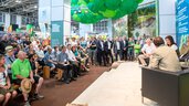 Großes Publikum verfolgt eine Podiumsdiskussion an einem Messestand mit Bühne, Sitzgelegenheiten und grüner Deckendekoration.