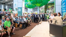 Großes Publikum verfolgt eine Podiumsdiskussion an einem Messestand mit Bühne, Sitzgelegenheiten und grüner Deckendekoration.