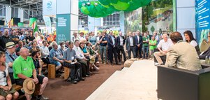  Großes Publikum verfolgt eine Podiumsdiskussion an einem Messestand mit Bühne, Sitzgelegenheiten und grüner Deckendekoration.
