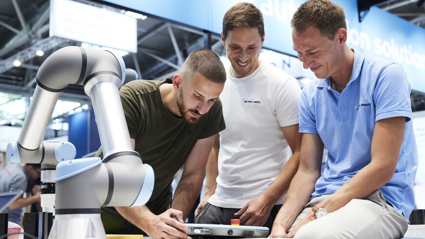 Three men interacting at a booth with a tablet to remotely control the robotic arm next to them.