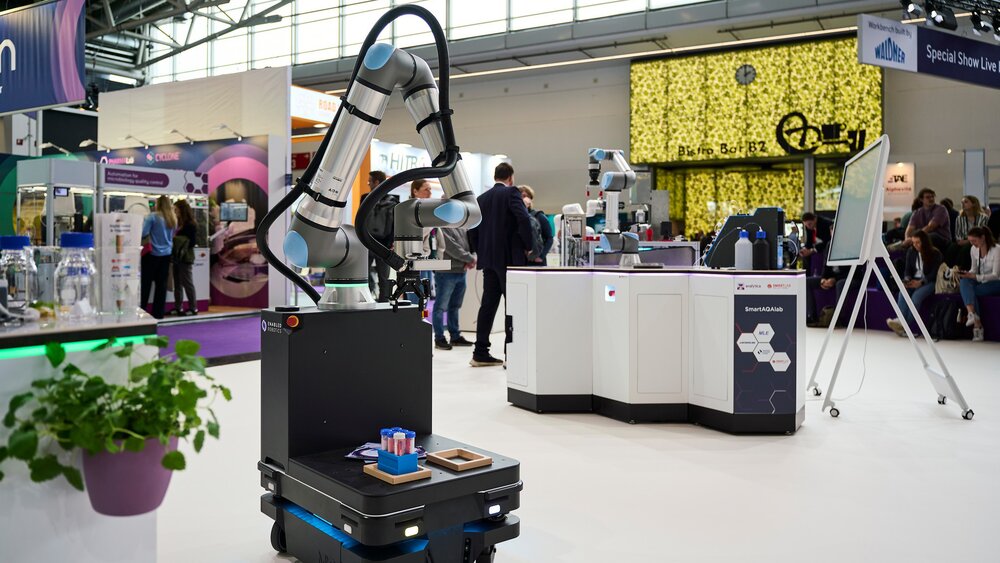 A mobile robotic arm holding laboratory samples at a stand in a modern exhibition hall.