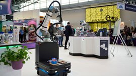A mobile robotic arm holding laboratory samples at a stand in a modern exhibition hall.