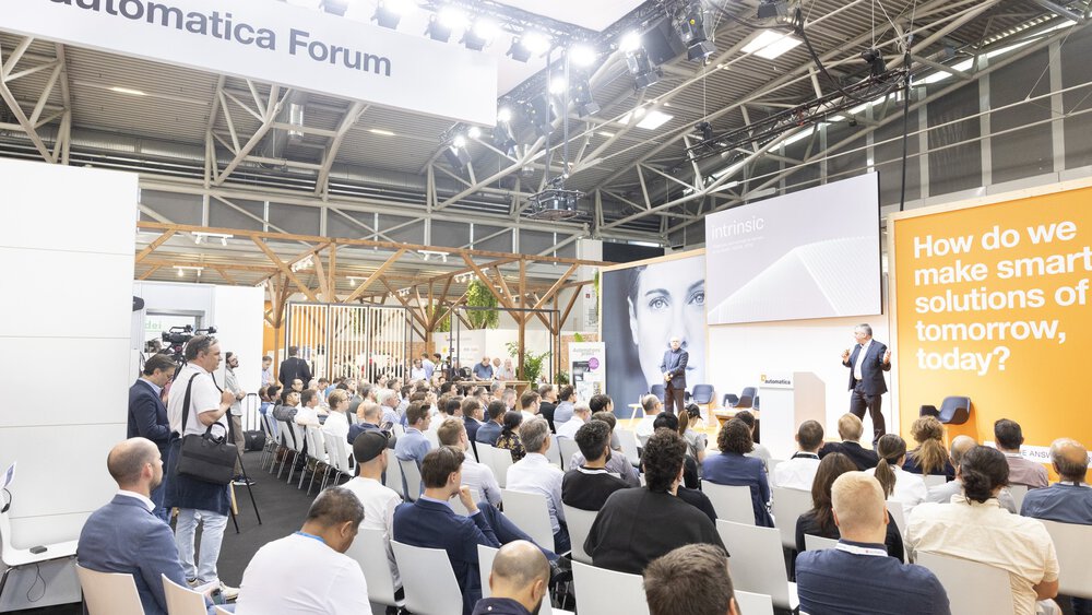 The audience listens to a speaker in the Automatica Forum, with a discussion topic displayed on a large screen.