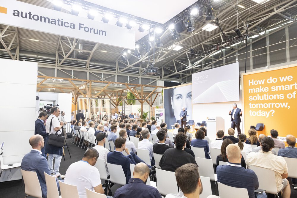The audience listens to a speaker in the Automatica Forum, with a discussion topic displayed on a large screen.