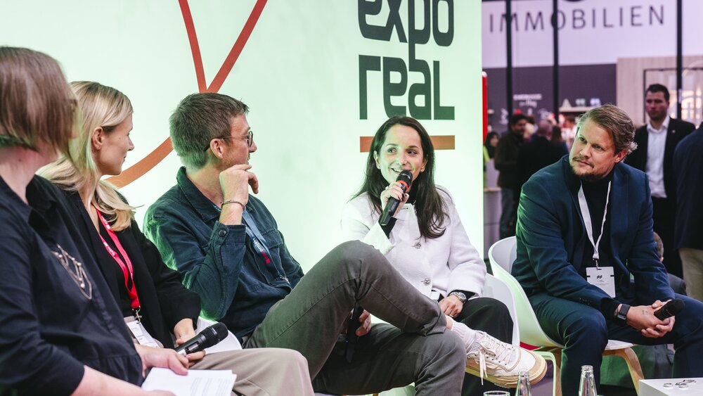 A group of five panelists sits on a stage at Expo Real. One woman speaks into a microphone while the others listen attentively. The Expo Real logo is visible behind them. In the background, trade fair visitors and various booths can be seen in the brightly lit hall.
