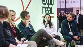 A group of five panelists sits on a stage at Expo Real. One woman speaks into a microphone while the others listen attentively. The Expo Real logo is visible behind them. In the background, trade fair visitors and various booths can be seen in the brightly lit hall.