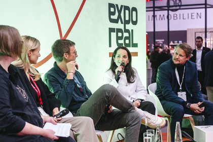 Eine Gruppe von fünf Panelteilnehmenden sitzt auf einer Bühne der Expo Real. Eine Frau mit Mikrofon spricht, während die anderen aufmerksam zuhören. Hinter ihnen ist das Expo-Real-Logo zu sehen. Im Hintergrund stehen Messebesucher und verschiedene Messestände in einer hell beleuchteten Halle.