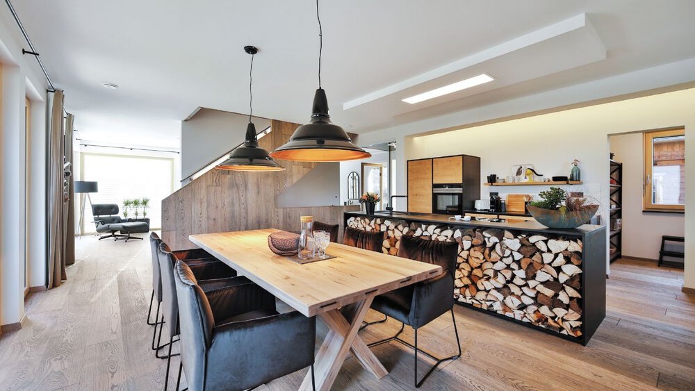Moderne Küche mit einem Esstisch aus Holz, schwarzen Stühlen, Hängelampen und einem Lager für Brennholz unter der Arbeitsplatte.