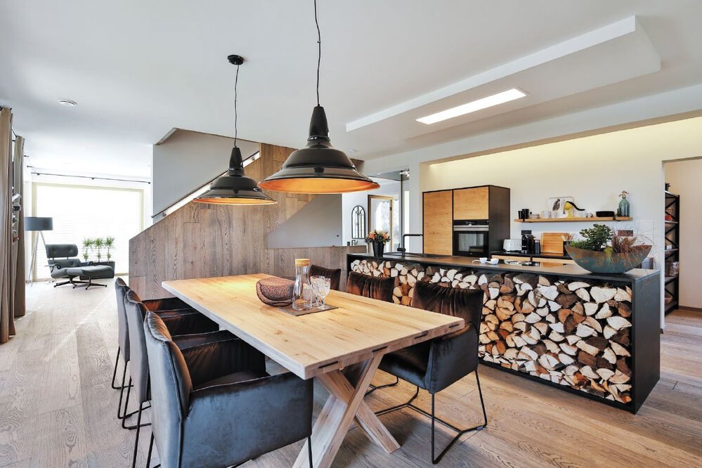 Moderne Küche mit einem Esstisch aus Holz, schwarzen Stühlen, Hängelampen und einem Lager für Brennholz unter der Arbeitsplatte.