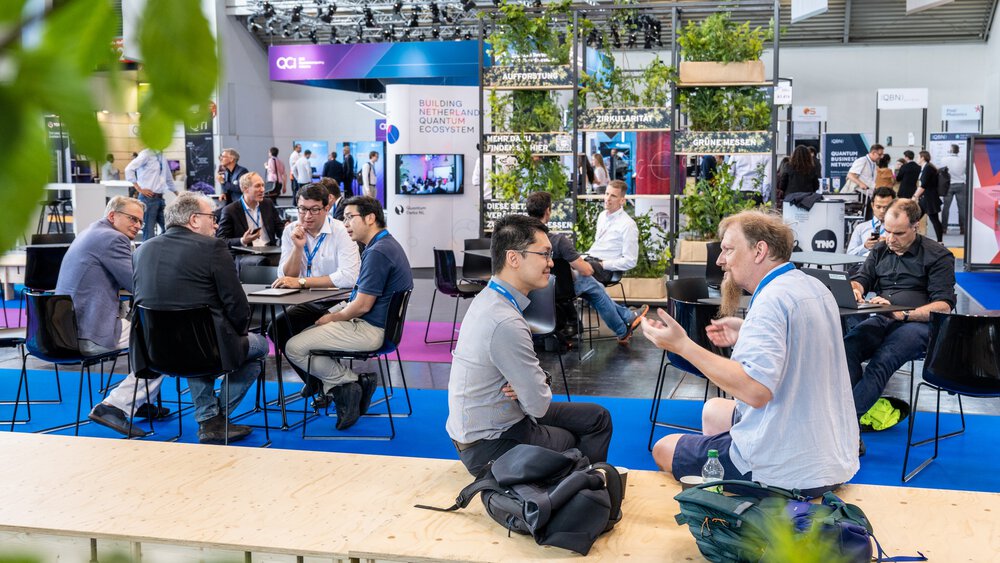 People sit and discuss in a modern conference area with a blue floor and green plants and immerse themselves in the world of quantum technology.