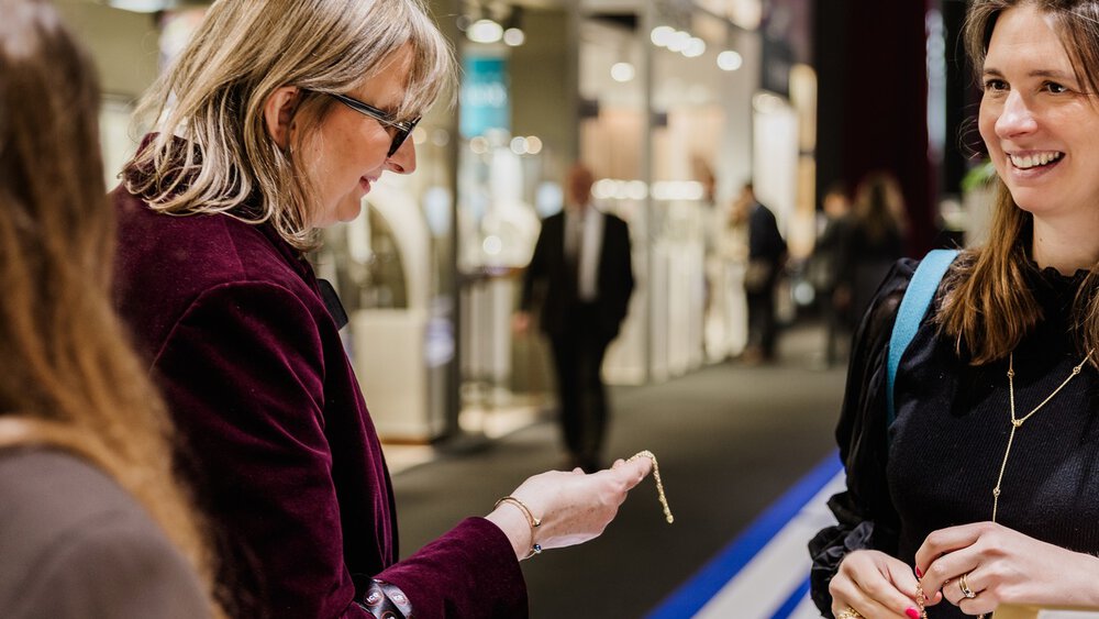 Two women smile and examine jewellery in a bright, modern exhibition hall.